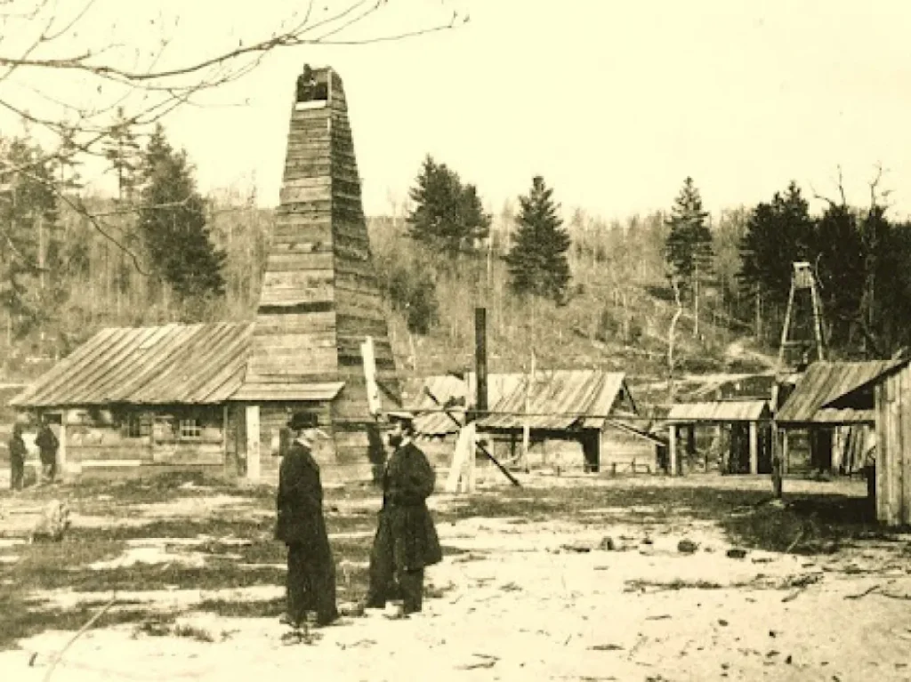 Sepia-toned photo of an early oil drilling site with a wooden derrick, several wooden buildings, and a few people in period clothing standing in the foreground. Trees and rolling hills are in the background.