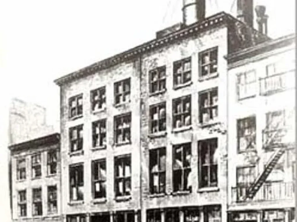 A black-and-white photo of an old multi-story factory building with many windows, some open. A fire escape is attached to the side, and there are water tanks and chimneys on the roof.