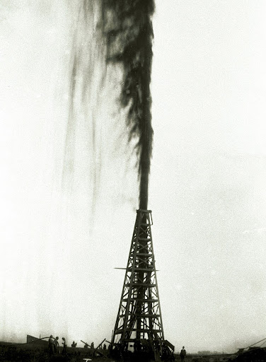 A tall oil derrick gushes a large stream of dark oil high into the air, with several small figures of people standing nearby, silhouetted against a pale sky.