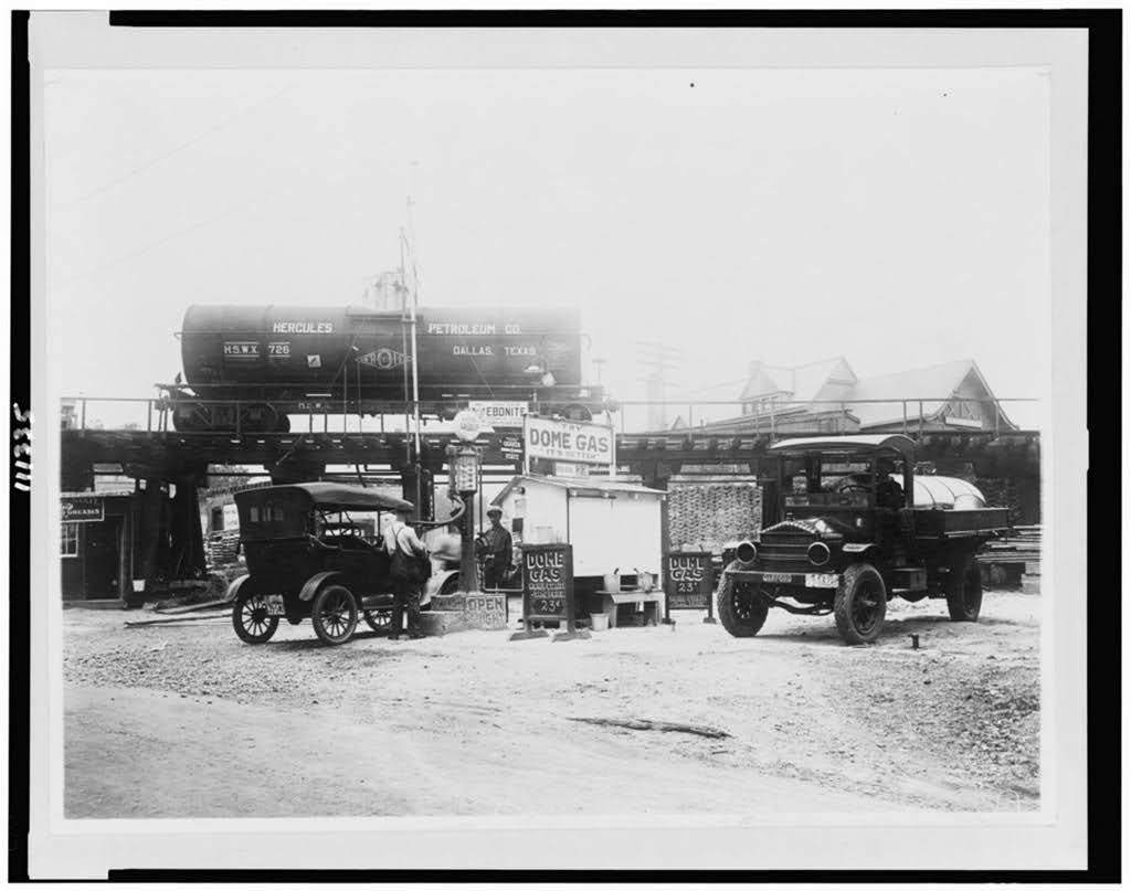 Historic black-and-white photo of a small gas station with two vintage vehicles parked; a fuel tank labeled “Hercules Petroleum Co.” sits on a raised platform behind the station. Signs read “Dome Gas” and offer oil and gas for sale.