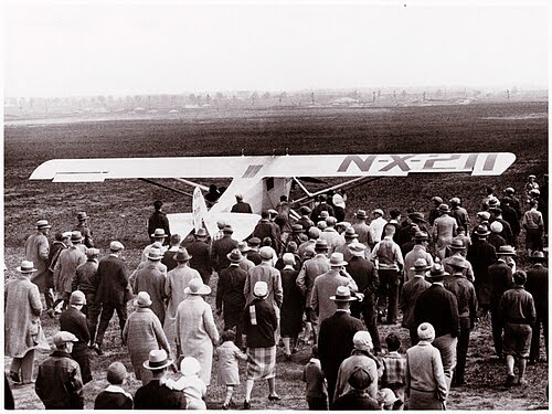 A large crowd gathers around a small white monoplane with "NX-211" on its wing, standing on a grassy field. Most people wear hats and coats, suggesting the scene is from the early 20th century.