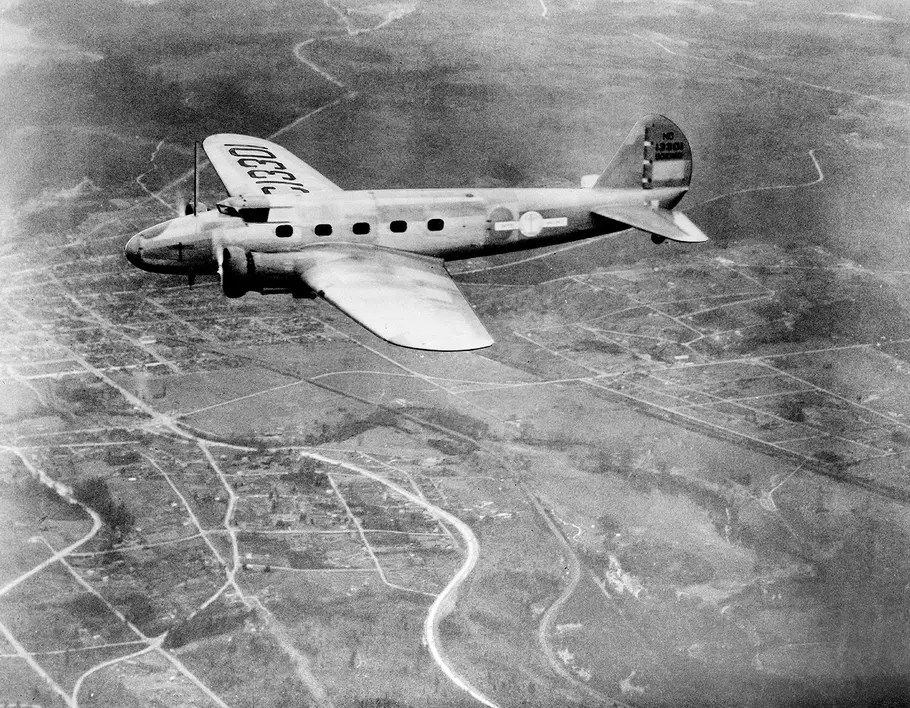 A vintage black and white photo of a military airplane flying above a landscape with visible roads and fields below. The aircraft has a U.S. insignia and markings on its wing and tail.