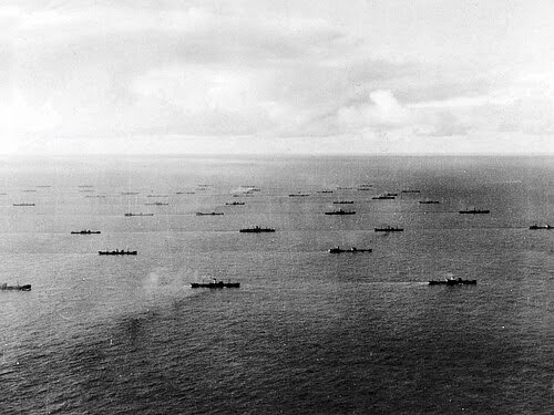 A large fleet of ships, widely spaced, sails across a calm ocean under a mostly cloudy sky; the vessels are spread out in several rows as far as the horizon.