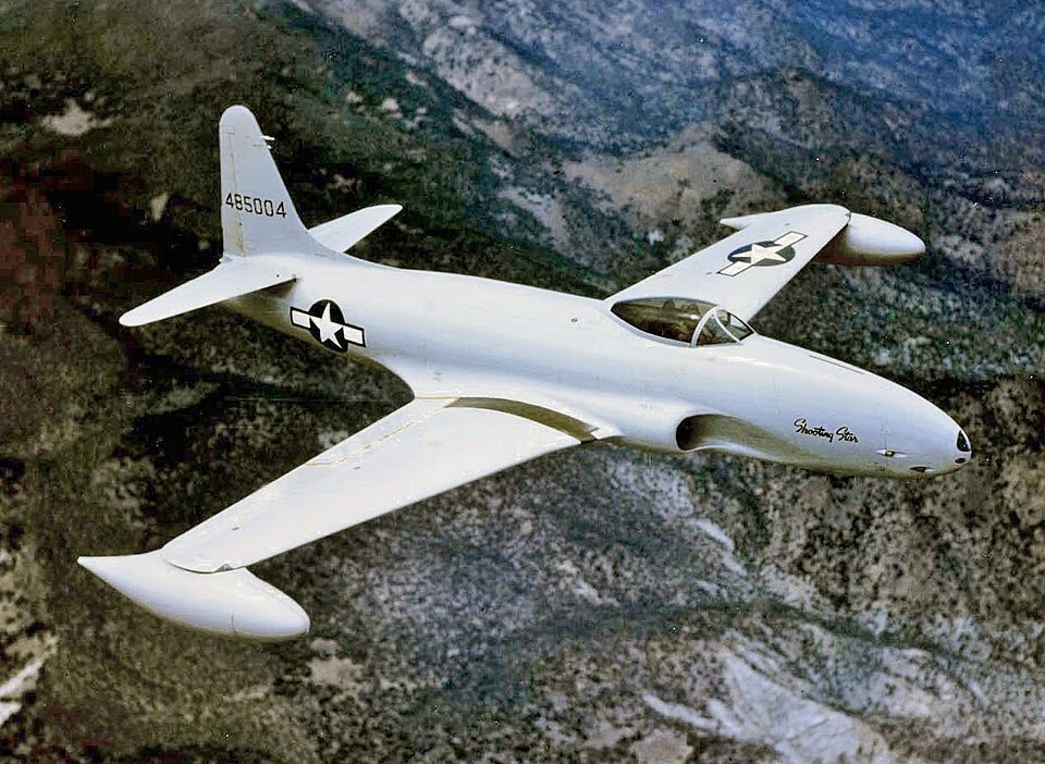 A white military jet with U.S. Air Force markings flies over a mountainous landscape. The aircraft has a sleek, streamlined design and "Shooting Star" written on the nose.