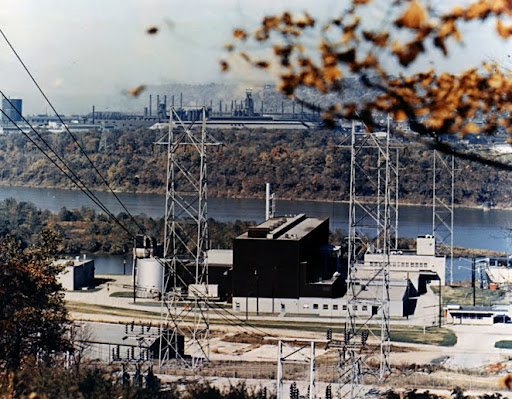 A power plant with tall metal towers and electrical infrastructure sits near a river, surrounded by trees and industrial buildings in the distance; autumn leaves partially frame the scene.
