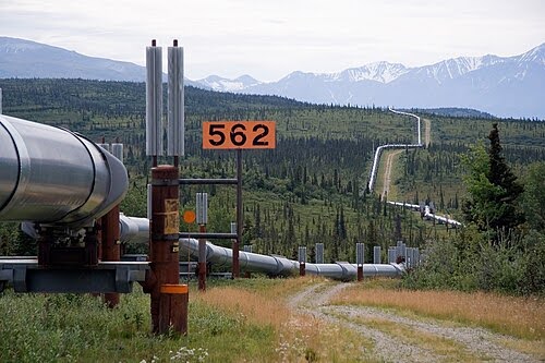 A large pipeline runs through a green, hilly landscape with mountains in the background. A sign marked “562” stands next to the pipeline, which curves into the distance. The scene is outdoors under a cloudy sky.