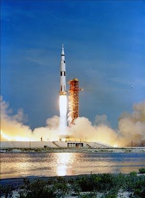 A large rocket launches from its pad, with bright flames and smoke billowing beneath it, against a blue sky. Water and greenery are visible in the foreground.