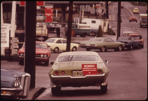 A green car with a large red "SORRY! TEMPORARILY OUT OF GAS" sign is parked on a street lined with other vehicles, near gas stations displaying Shell and Chevron signs. The sky is overcast.
