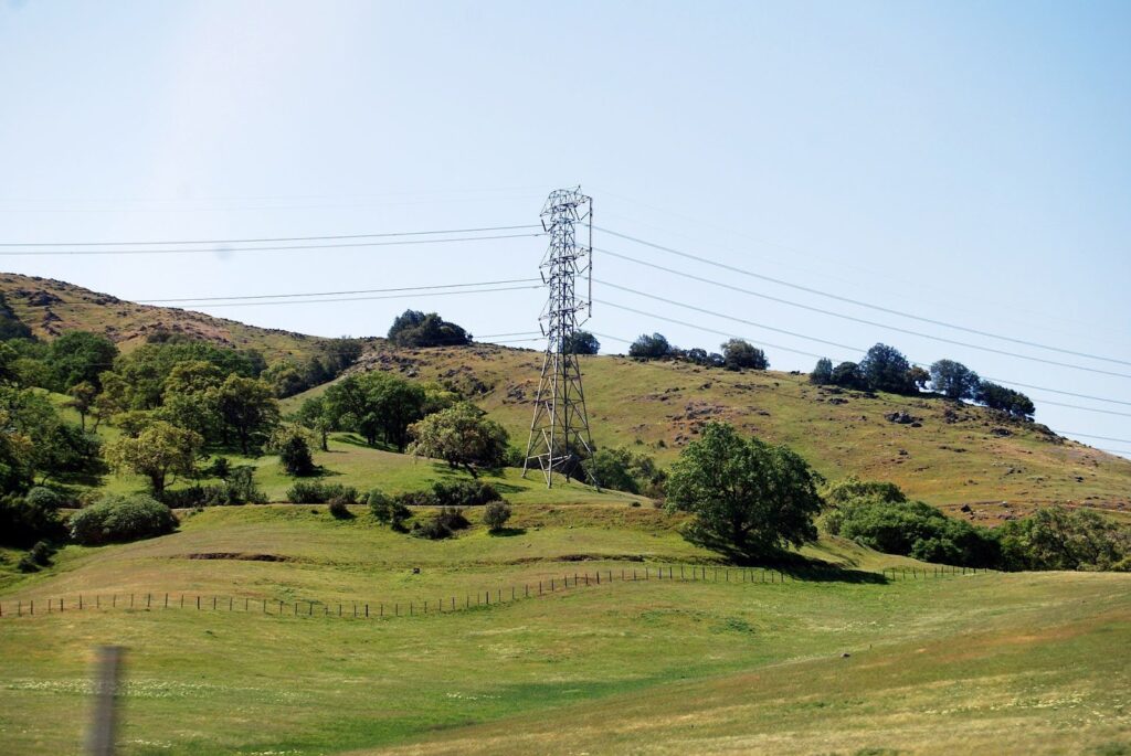 A large power line tower stands on a grassy hillside with scattered trees and a clear blue sky in the background. A fence runs along the green landscape in the foreground.