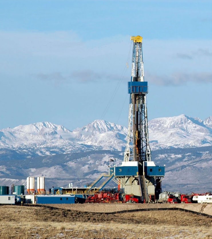 A large oil drilling rig stands in a grassy field with snow-capped mountains in the background. Several storage tanks, buildings, and equipment surround the rig under a clear sky.