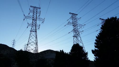 Tall electricity pylons and power lines stretch across a hilly landscape at dusk, with trees and silhouetted hills visible against the evening sky.