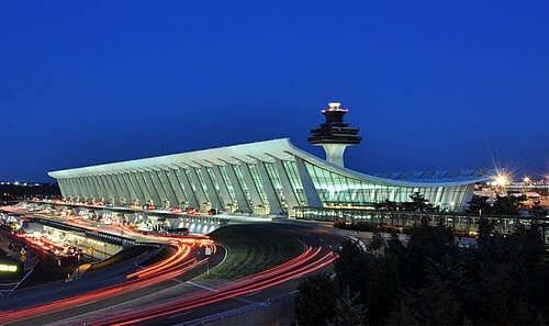 A modern airport terminal with a sweeping, curved roof and illuminated windows at dusk; light trails from moving vehicles are visible in the foreground.