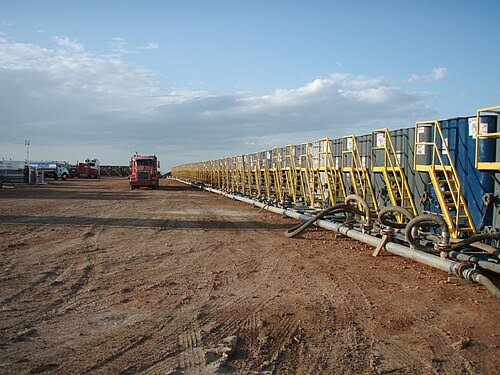 A row of yellow and blue industrial equipment, likely used for oil or gas extraction, is lined up on a dirt lot under a blue sky with scattered clouds; a red truck is parked nearby.