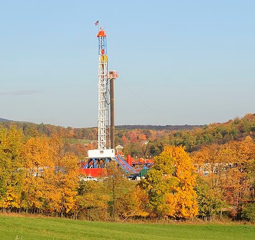 A drilling rig stands among colorful autumn trees in a rural landscape under a clear blue sky, with distant hills visible in the background.