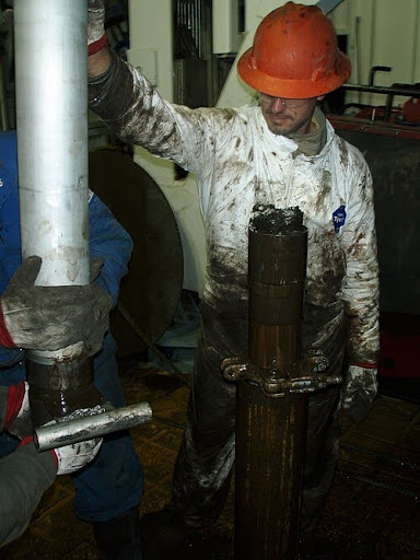 A worker in a dirty orange hard hat and oil-stained coveralls handles a large, muddy metal pipe on an industrial worksite, while another gloved worker assists.