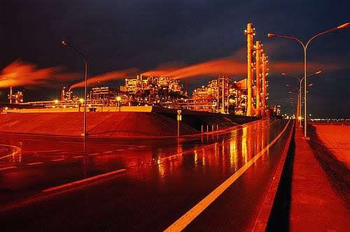 A brightly lit industrial plant at night emits steam or smoke, reflecting orange and yellow lights on the wet road and surrounding area under a dark sky. Tall streetlights line the roadway.