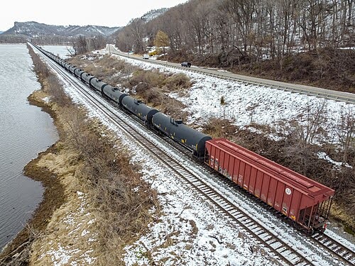 A long freight train with black tanker cars and a single red boxcar travels along snowy tracks beside a river, with a road and leafless trees on the right and hills in the background.