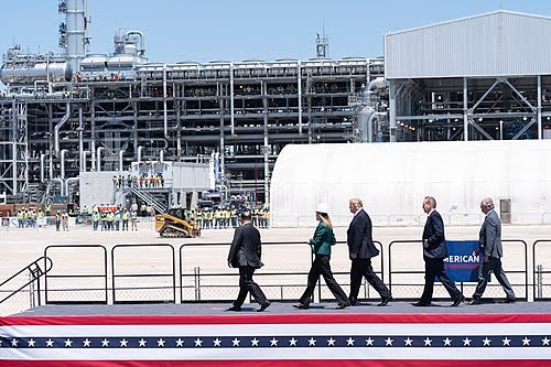 Five people in business attire walk in a line outside an industrial facility with metal pipes and structures, while workers stand in the background. A stage with an American flag pattern is in the foreground.