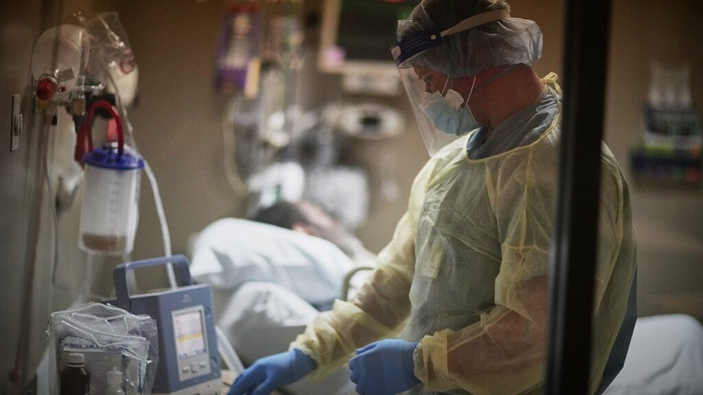 A healthcare worker in protective gear and a face shield attends to a patient lying in a hospital bed, adjusting medical equipment in a dimly lit hospital room.