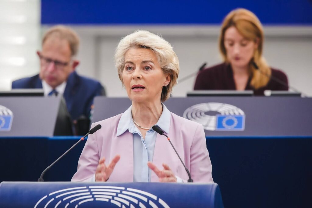 A woman speaks at a podium in a formal setting, with two people seated behind her and European Parliament symbols visible on the blue panels.