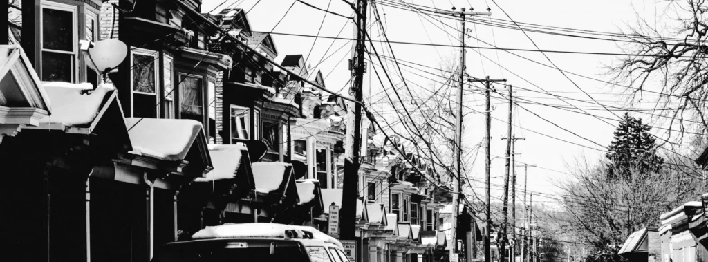 A black and white photo of a row of attached houses with snow on the roofs, many power lines overhead, and cars parked along the street; bare trees are visible in the background.