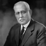 An older man with short gray hair, glasses, and a mustache, wearing a dark suit, vest, and tie, sits in front of a blurred dark background, looking slightly to the side with a serious expression.