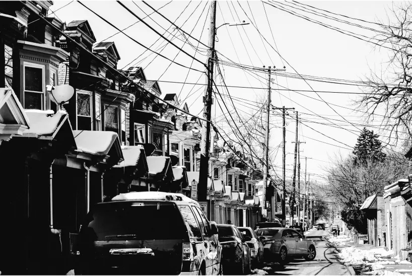 Black and white photo of a narrow city street lined with parked cars and row houses, with numerous overhead utility wires running between tall poles. Snow patches are visible on rooftops and on the ground.