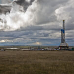 An oil drilling rig stands on open grassland under a dramatic, cloudy sky. Several colorful storage tanks are nearby, with a few vehicles and equipment scattered around the site.