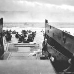 Soldiers disembark from a landing craft into the water, heading toward a beach under heavy smoke and gunfire during a military invasion, with more troops and equipment visible in the distance.