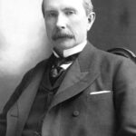 A black-and-white portrait of a man in a suit with a mustache, seated and looking slightly to the side. He has short hair and wears a vest, tie, and pocket watch.