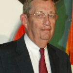 A man in a dark suit, white shirt, and red patterned tie stands in front of a colorful abstract background. He has short hair, glasses, and a neutral expression.