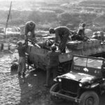 Soldiers load supplies onto a military vehicle while others stand nearby; a jeep marked “G-1” is parked in the foreground. Stacks of crates and rough terrain are visible in the background.