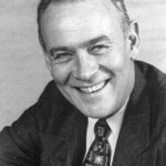 Black and white headshot of a smiling middle-aged man wearing a suit and patterned tie.